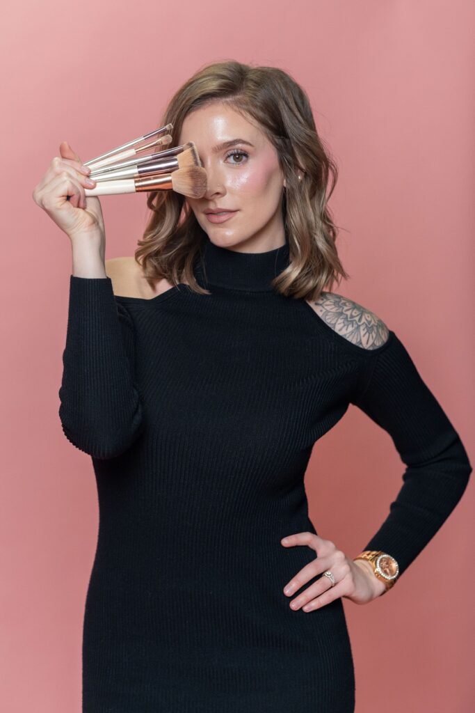 Alie Reino holding makeup brushes standing up against a pink background