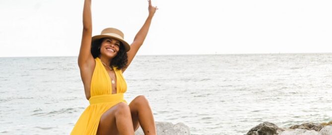 Tiffiny sitting on rocks on the beach in a yellow dress with white hat.